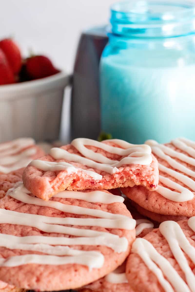 a partially eaten strawberry cookie on a stack of cookies with a cup of milk.