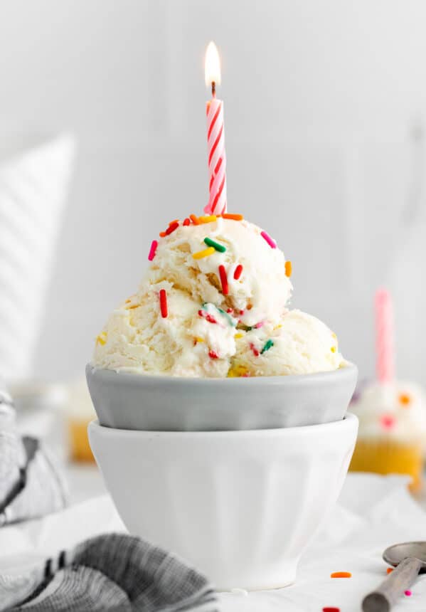 side view of 3 scoops of birthday cake ice cream in a gray bowl set in a white bowl topped with a lit pink birthday candle.