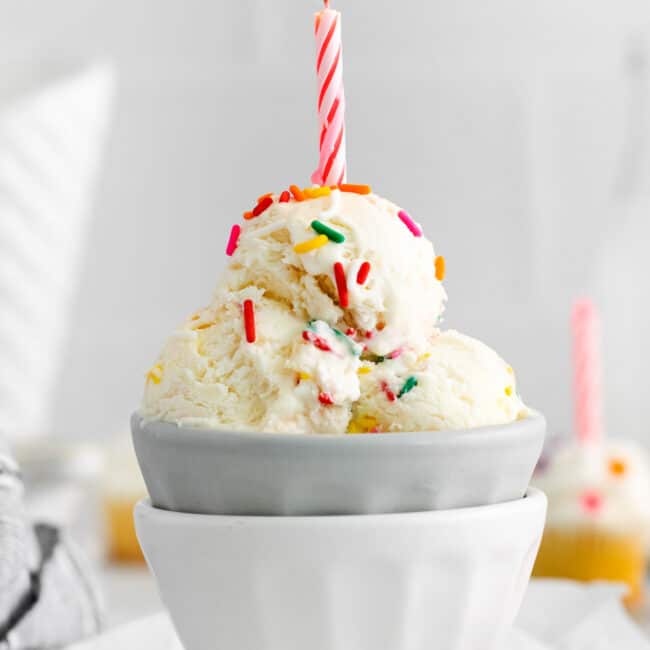 side view of 3 scoops of birthday cake ice cream in a gray bowl set in a white bowl topped with a lit pink birthday candle.