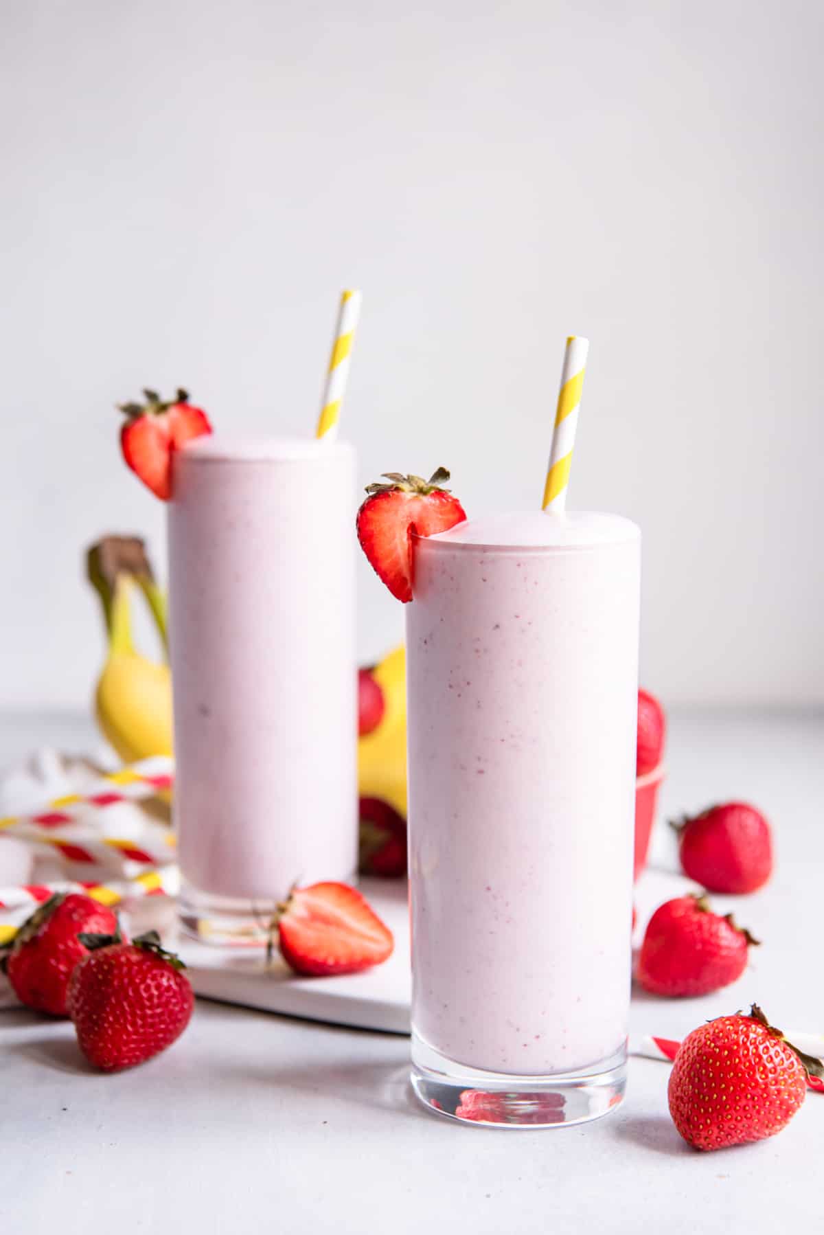 two strawberry banana smoothies in tall glasses, with yellow-striped straws, and sliced strawberries scattered around on the table.