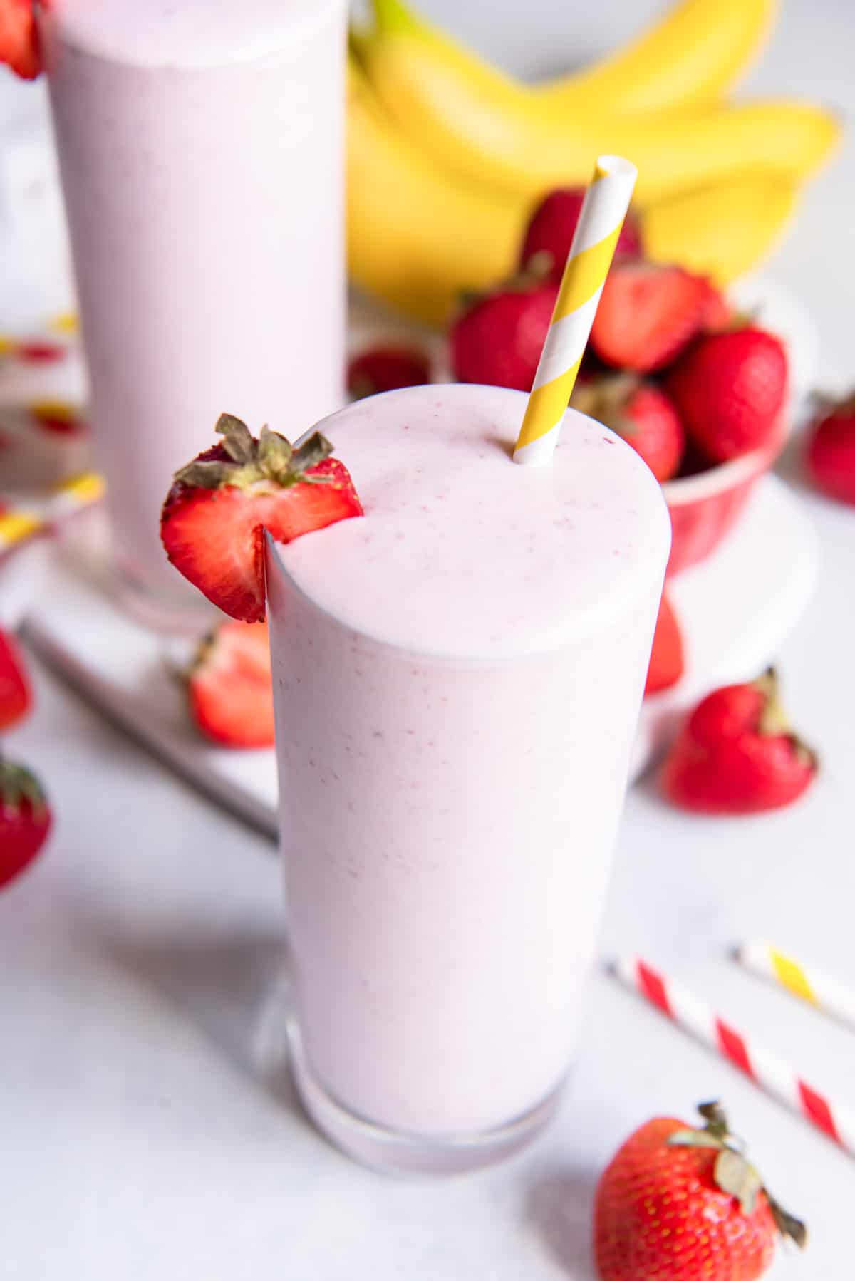 two strawberry and banana smoothies on a table top filled with strawberries and striped straws.