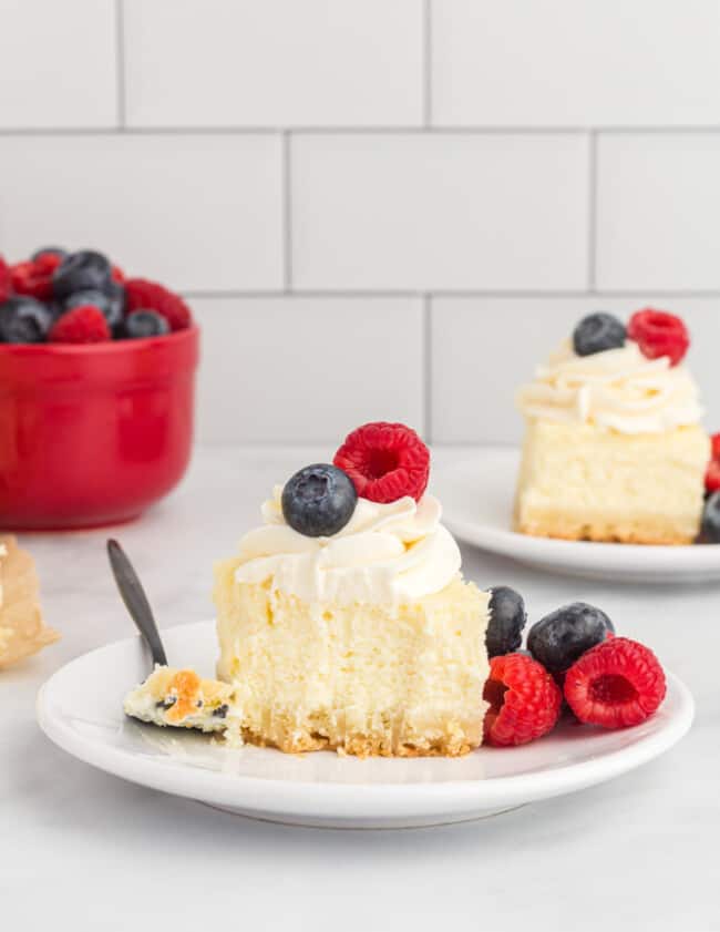 side view of a cheesecake bar on a white plate with berries, whipped cream, and a fork.
