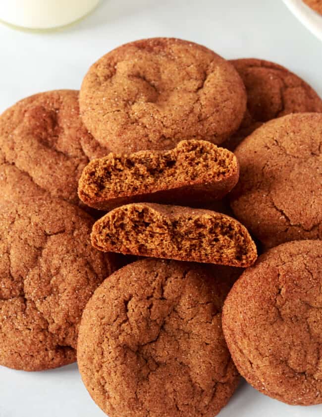 three-quarters view of gingersnap cookies in a pile on a white counter, one is halved and stacked on itself to show the interior texture.