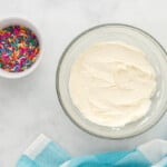 birthday cookie frosting in a white bowl next to a bowl of rainbow sprinkles.