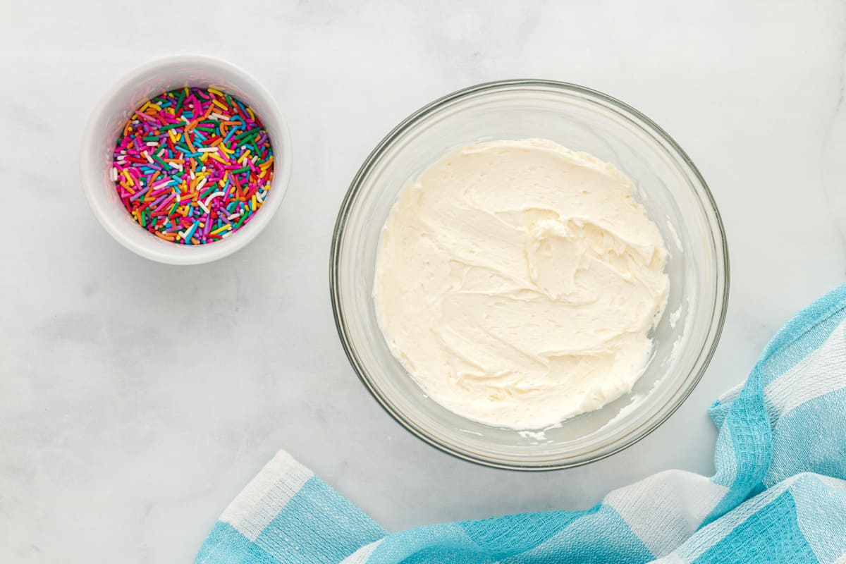 birthday cookie frosting in a white bowl next to a bowl of rainbow sprinkles.