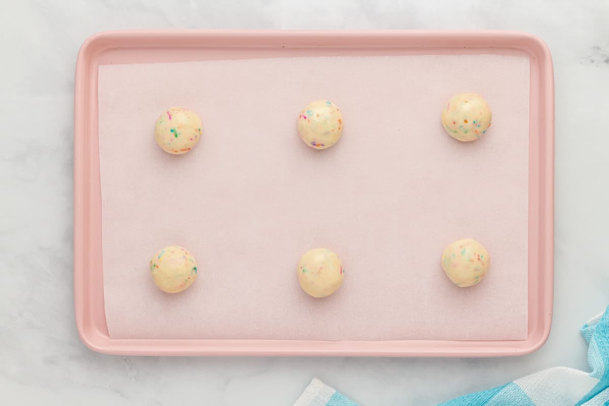 6 birthday cookie dough balls spaced out on a pink baking sheet.