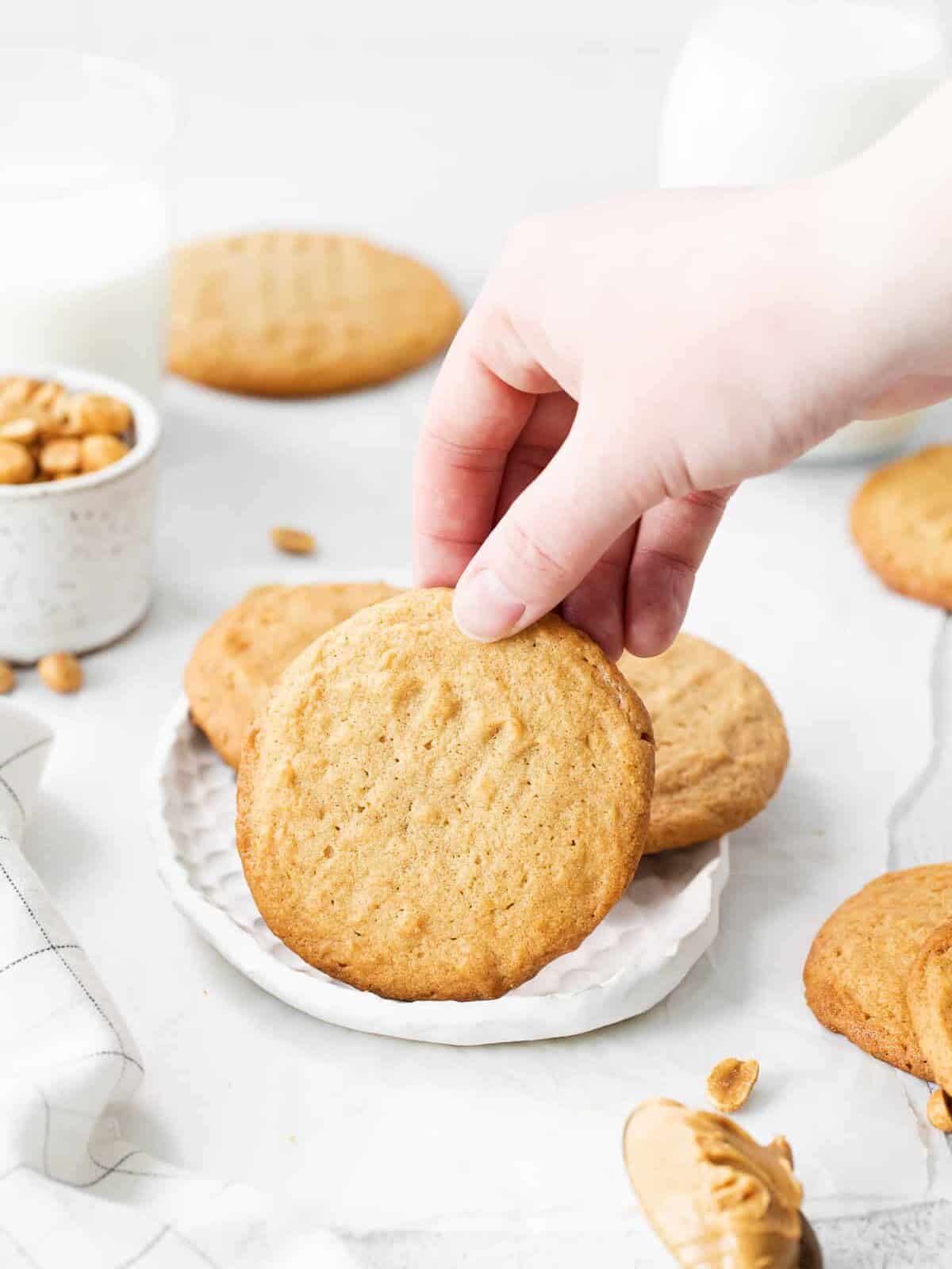 a hand grabbing 1 of 3 brown butter peanut butter cookies from a white plate.