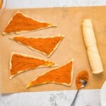 triangles of crescent roll dough are laid flat on a sheet of parchment paper, with orange pumpkin pie filling spread on top of each piece.