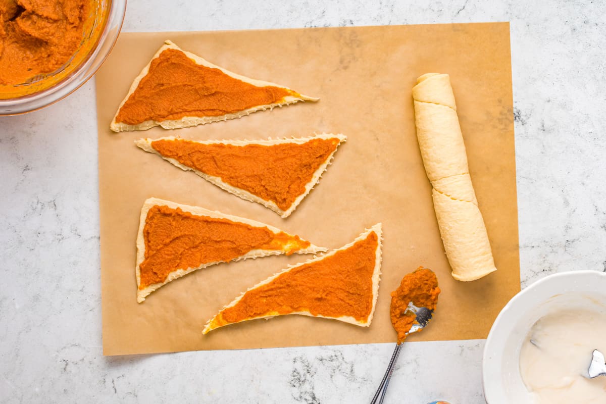 triangles of crescent roll dough are laid flat on a sheet of parchment paper, with orange pumpkin pie filling spread on top of each piece.