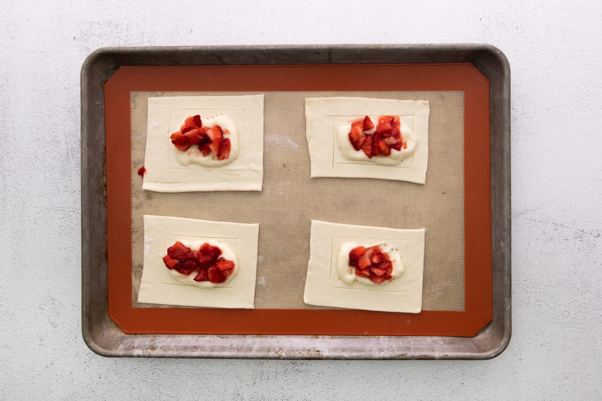 four squares of pastry with strawberries and cream on a baking sheet.
