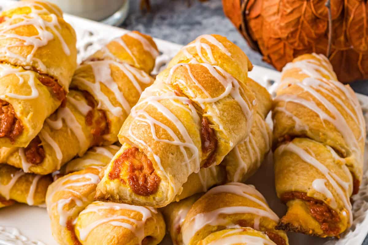 a serving tray of pumpkin pie crescent rolls is set on a table next to a glass of milk and a decorative orange pumpkin.