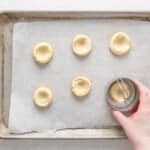 a hand using a biscuit cutter to round the edges of 6 baked dimpled shortbread cookies on a baking sheet.