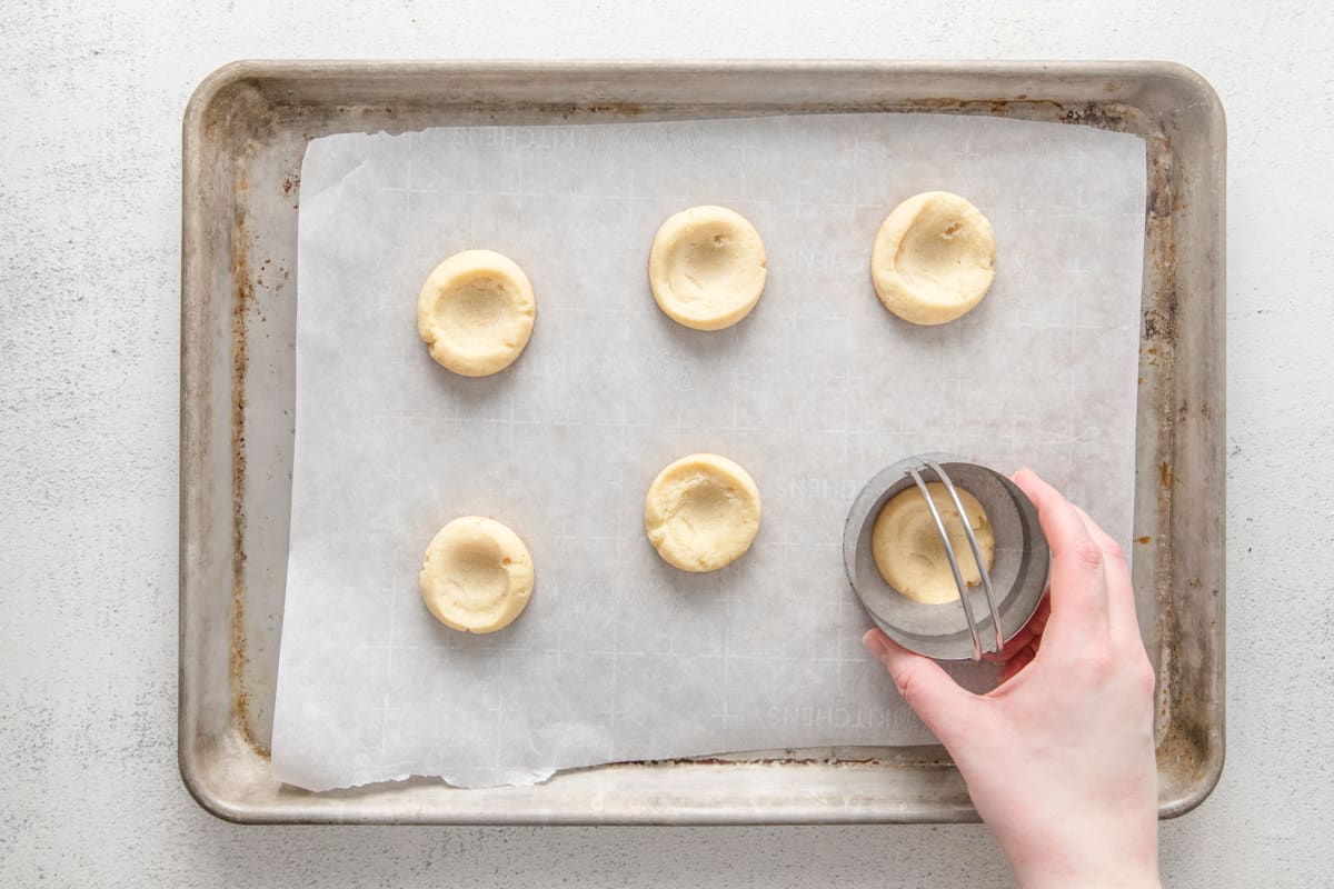 a hand using a biscuit cutter to round the edges of 6 baked dimpled shortbread cookies on a baking sheet.