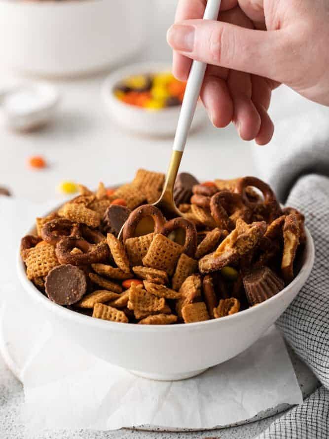 hand dipping a spoon into a bowl of a bowl of chocolate peanut butter snack mix