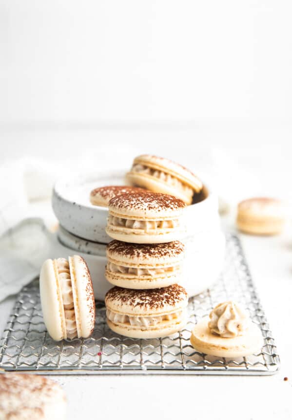 side view of 3 tiramisu macarons stacked in a tower next to a macaron on its side and a macaron half-filled with frosting in front of a white bowl filled with 3 macarons.