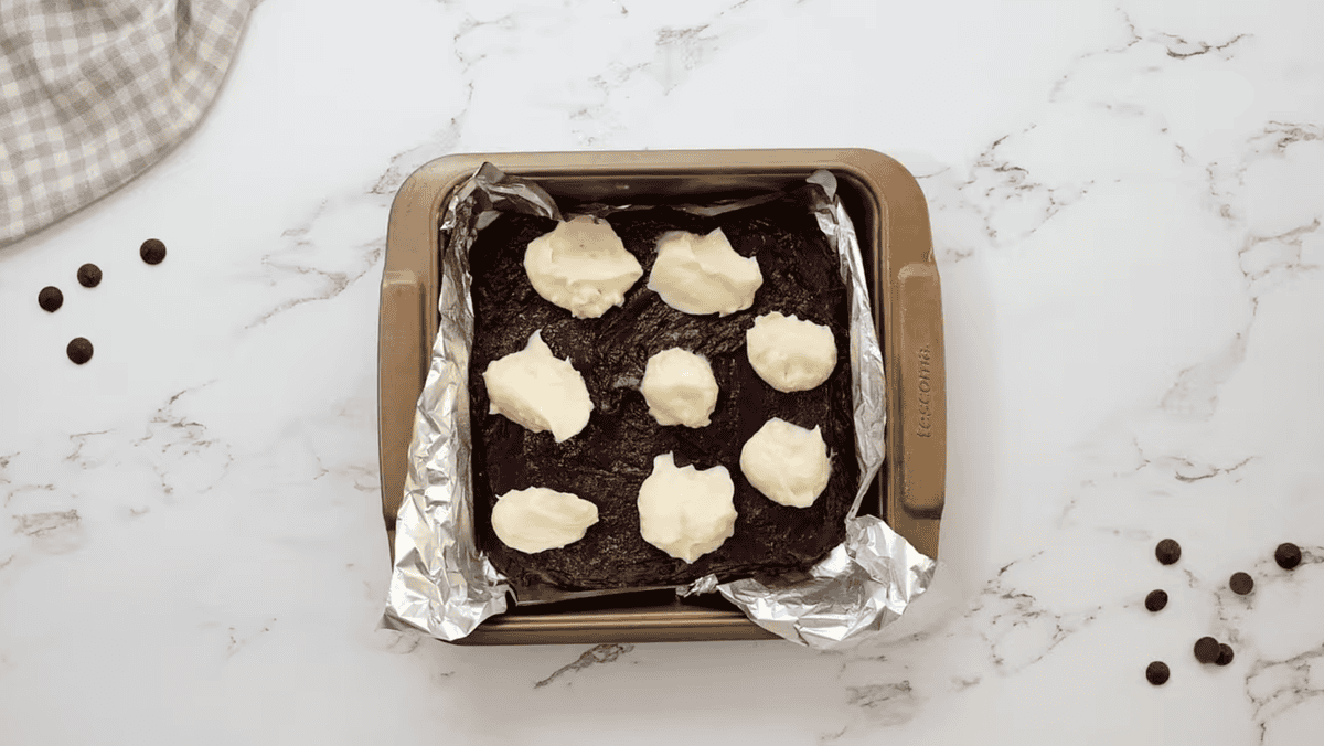 cream cheese dolloped over brownie batter in a foil lined baking pan.