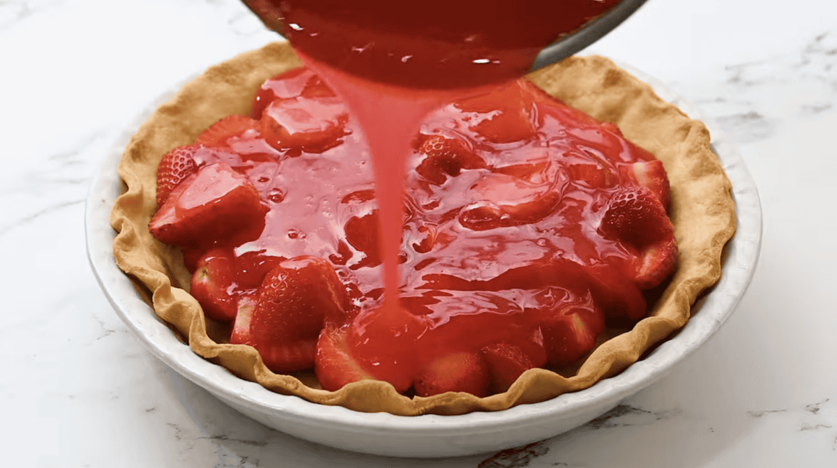 pouring red syrup over halved strawberries in a baked pie crust.