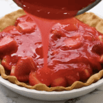 pouring red syrup over halved strawberries in a baked pie crust.