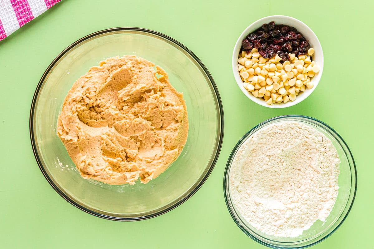 wet and dry ingredients in separate glass bowls next to a bowl of white chocolate and dried cranberries.