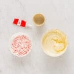 melted white chocolate in a bowl next to crushed candy canes, peppermint extract, and red food coloring.