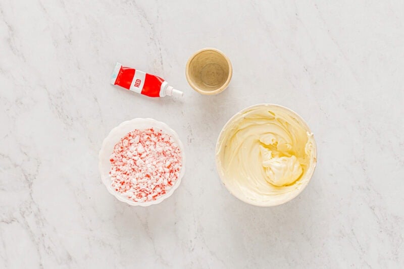 melted white chocolate in a bowl next to crushed candy canes, peppermint extract, and red food coloring.