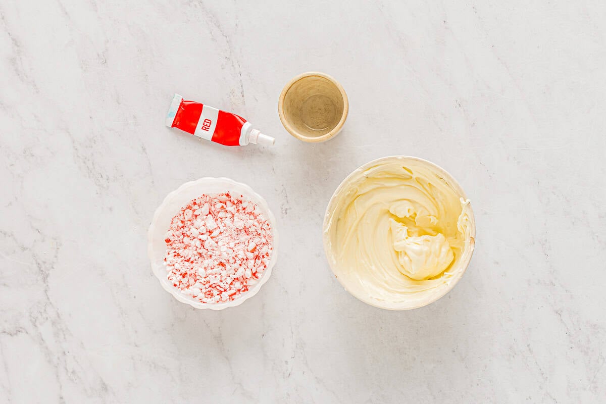 melted white chocolate in a bowl next to crushed candy canes, peppermint extract, and red food coloring.