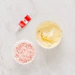 melted peppermint white chocolate in a bowl next to crushed candy canes and red food coloring.