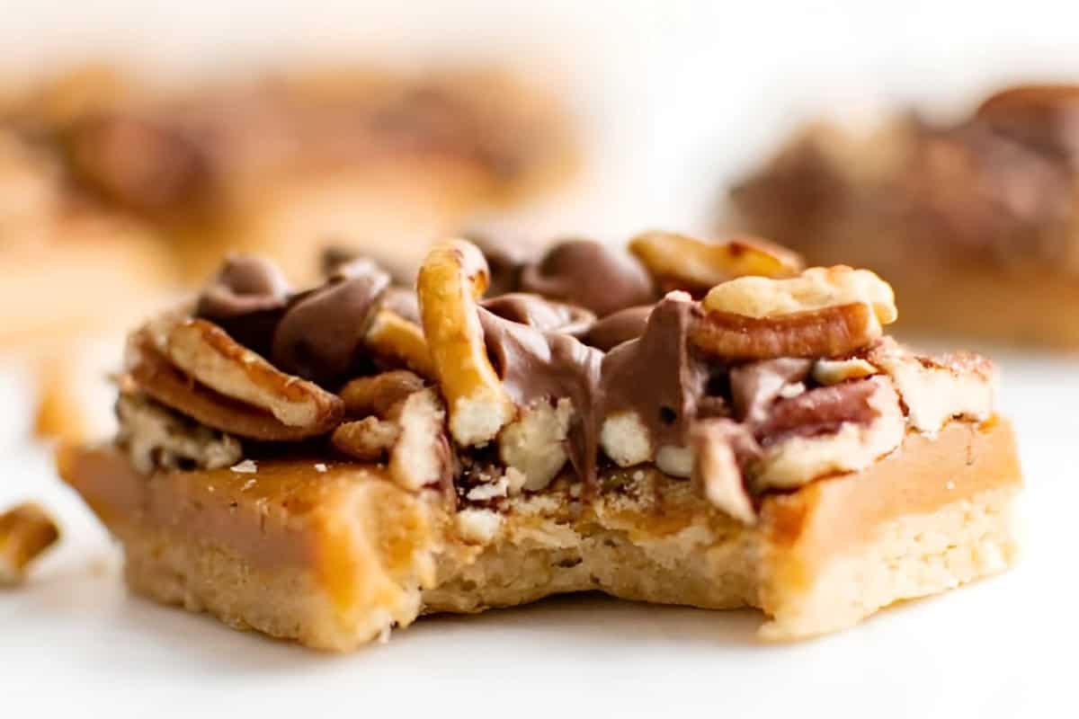 closeup side view of a bitten turtle bar, to show layers of shortbread, caramel, chocolate, and pecan.