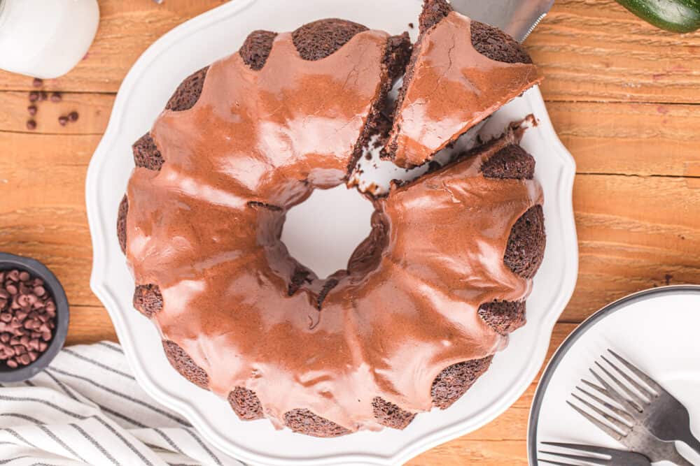 overhead view of glazed chocolate zucchini bundt cake on a white cake stand with a slice being removed by a cake server.