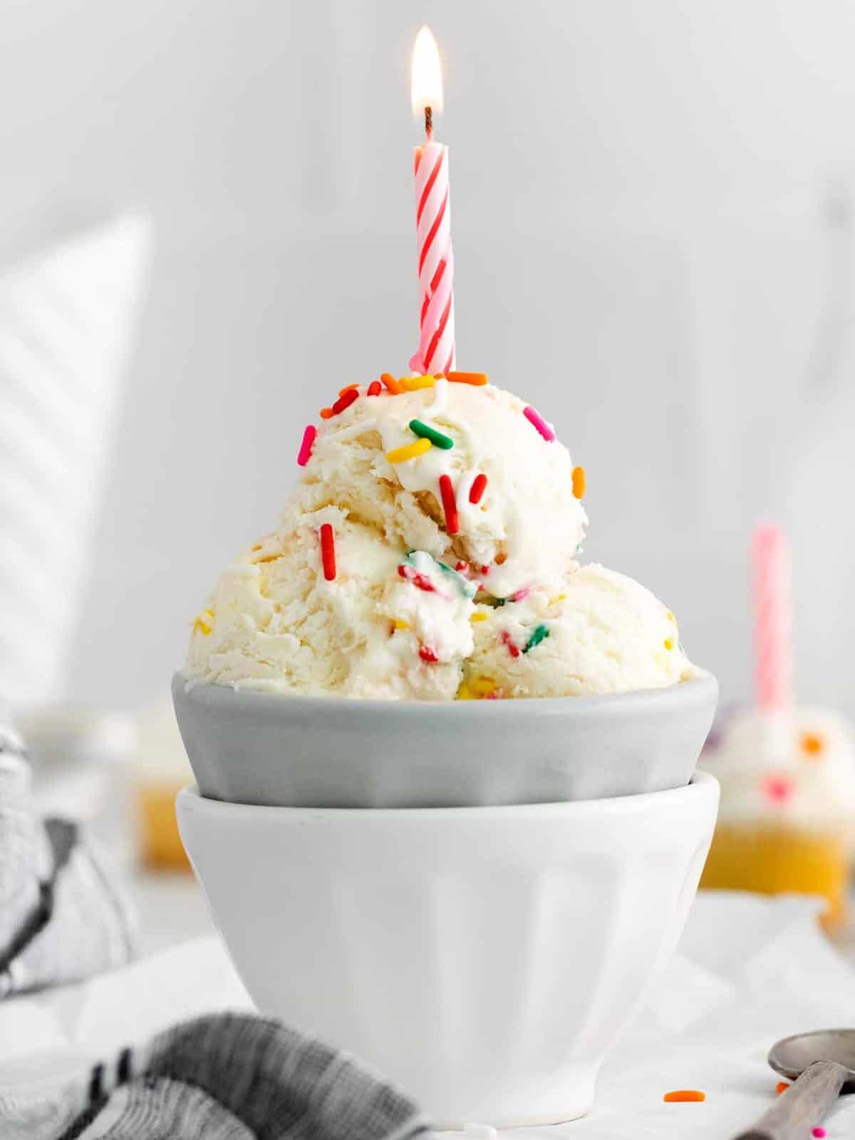 side view of 3 scoops of birthday cake ice cream in a gray bowl set in a white bowl topped with a lit pink birthday candle.