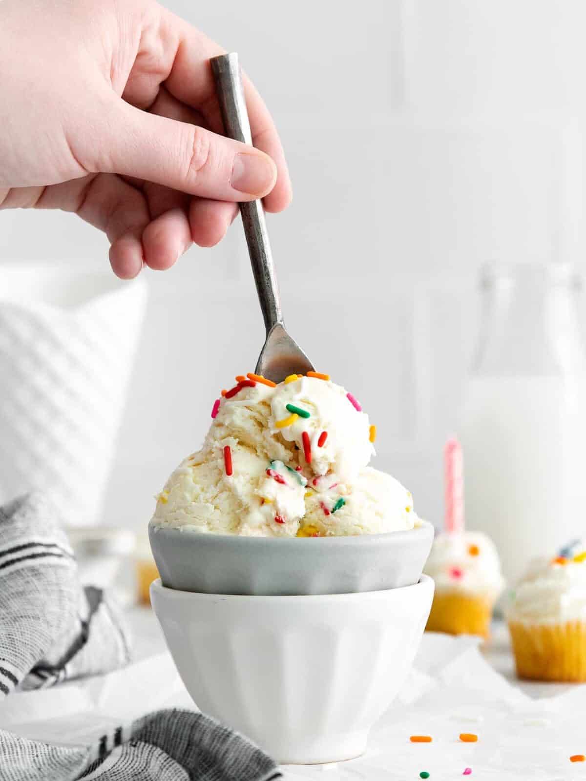 side view of a hand plunging a spoon into birthday cake ice cream in a gray bowl set in a white bowl.