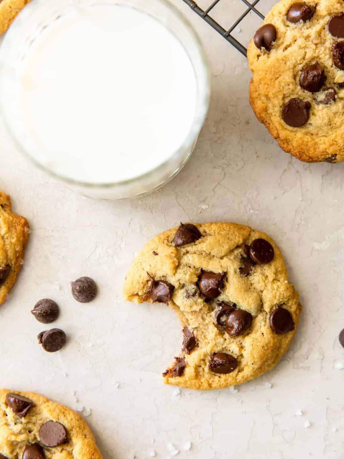 overhead view of a bitten gluten free chocolate chip cookie on a white table with a glass of milk.