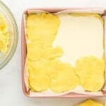 flat lemon cake mix dough pieces pressed over cheesecake filling in a square baking pan.