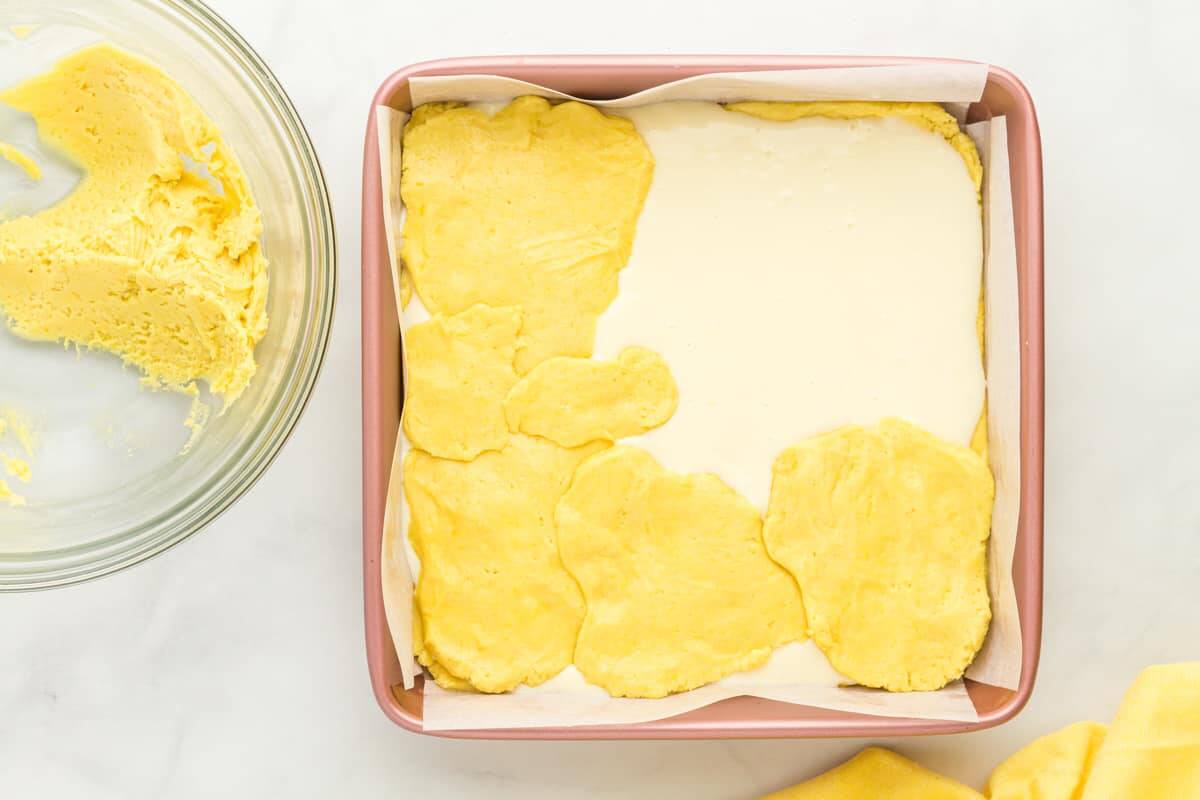flat lemon cake mix dough pieces pressed over cheesecake filling in a square baking pan.