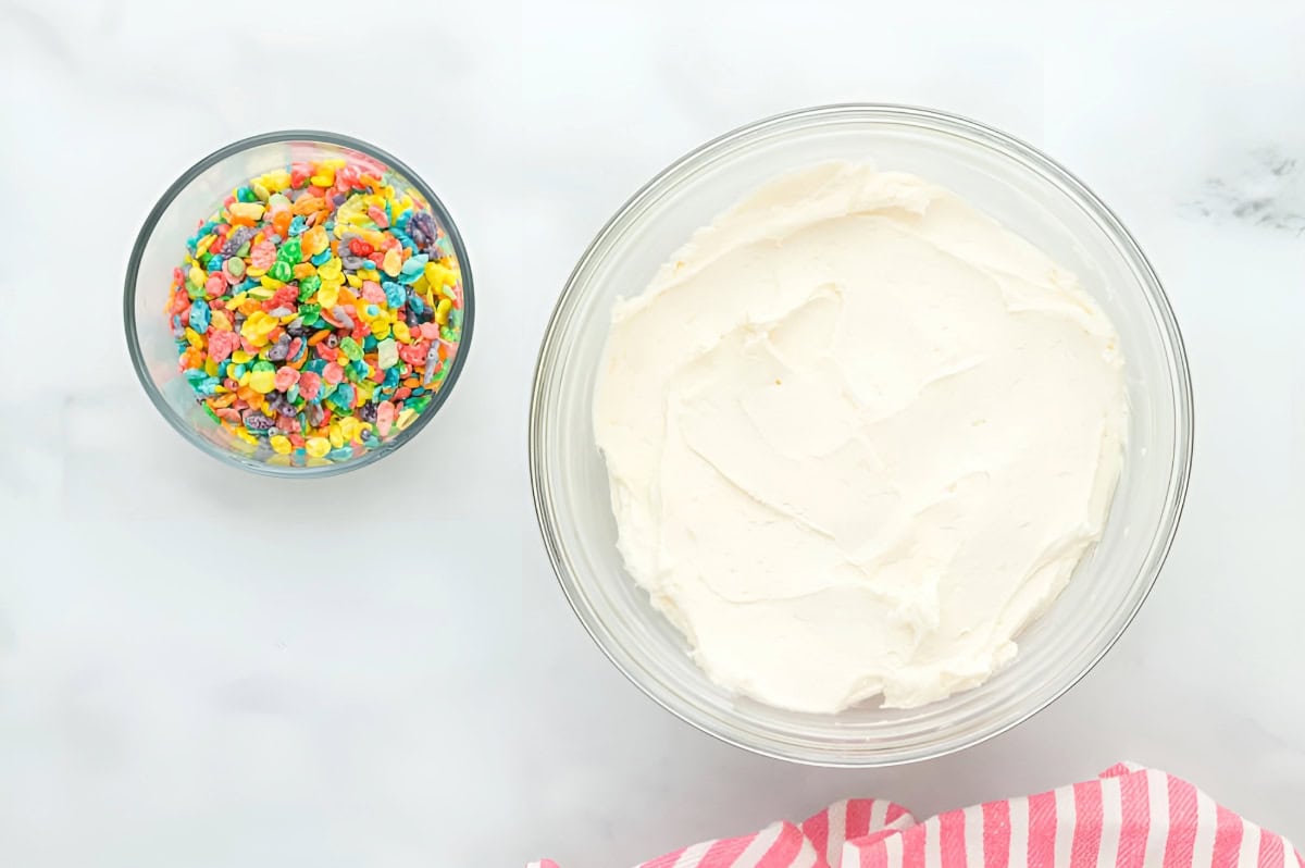 frosting in a glass bowl next to a dish of fruity pebbles.