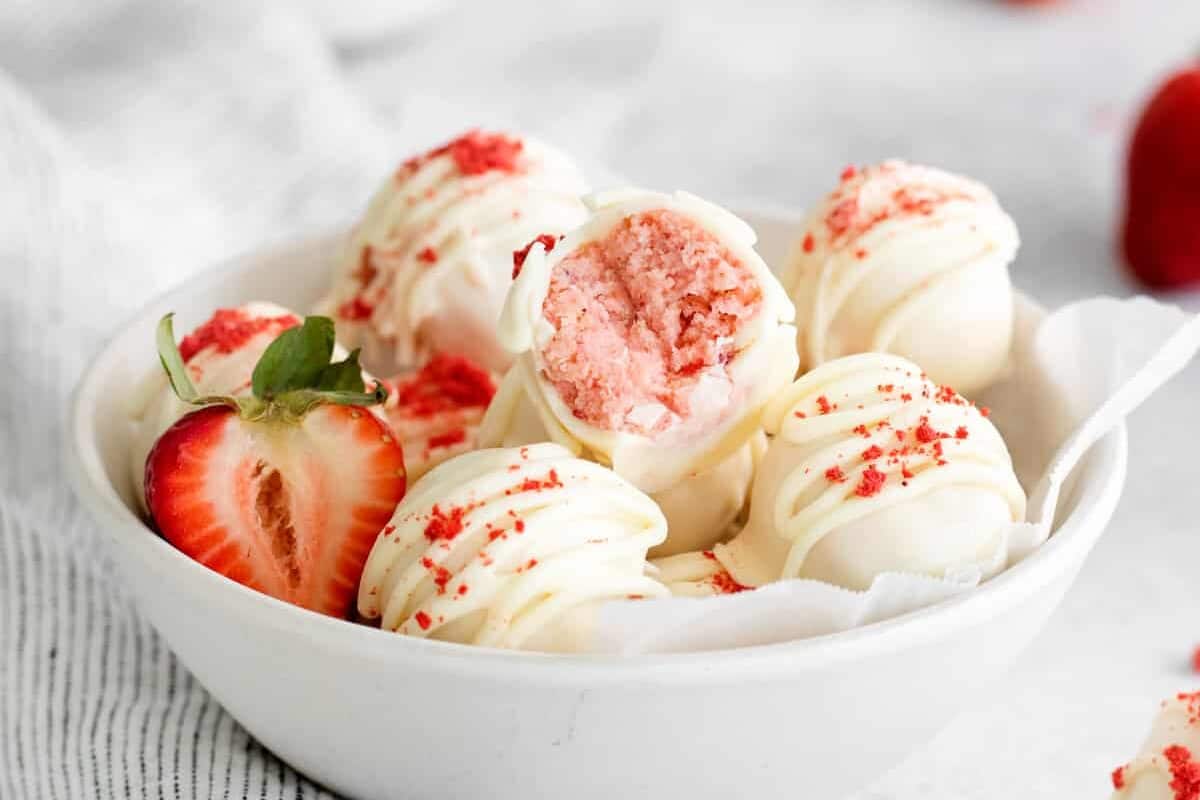 three-quarters view of white chocolate coated truffles in a white bowl, the top truffle has a bite taken out to reveal the strawberry cake at the center