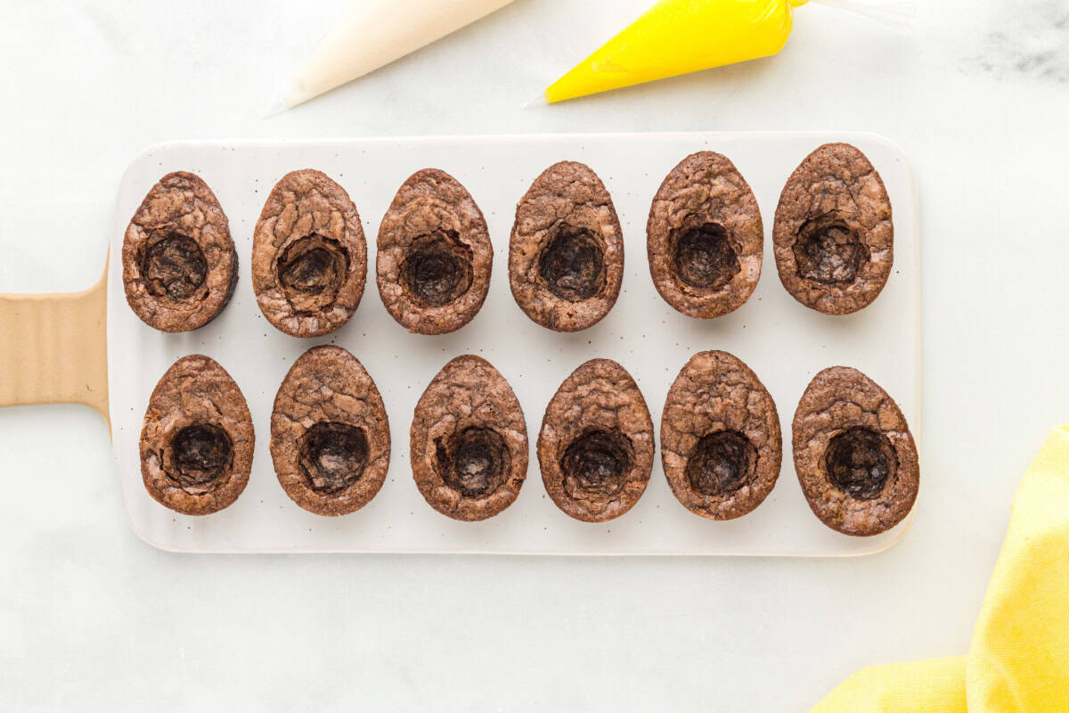 cooled egg-shaped brownies with hollowed-out yolk centers, surrounded by creme filling in piping bags.