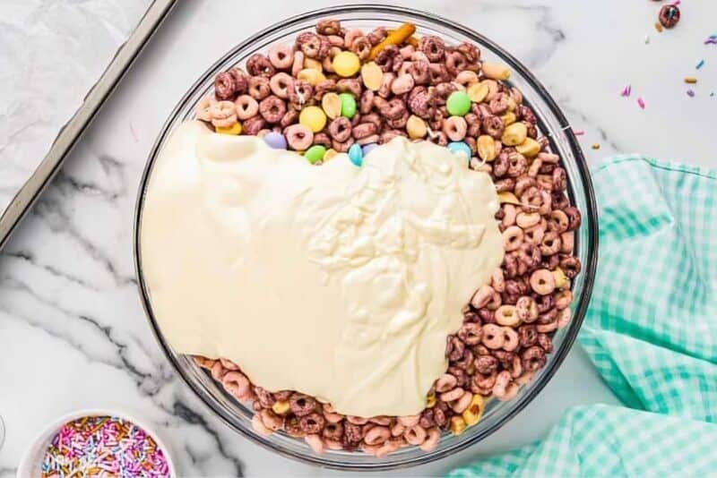 Melted white chocolate coating a bowl of snack mix ingredients.