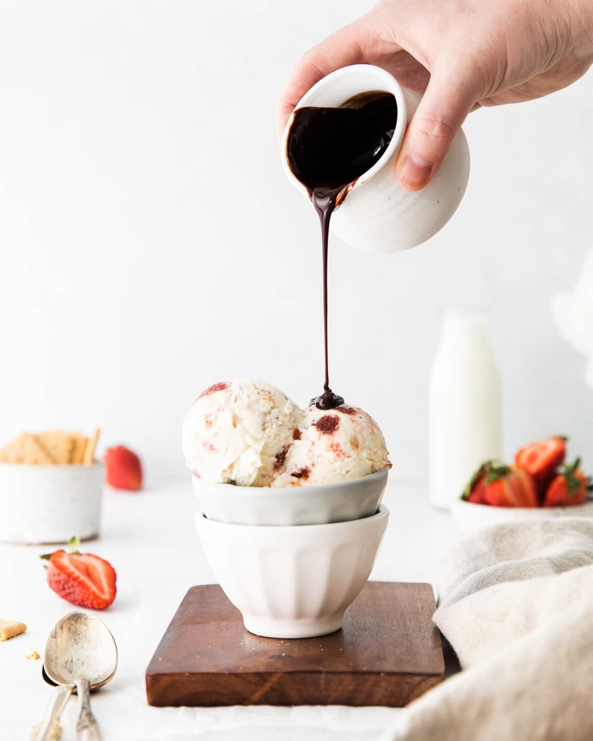 Pouring chocolate sauce over scoops of strawberry cream cheese ice cream in a bowl.
