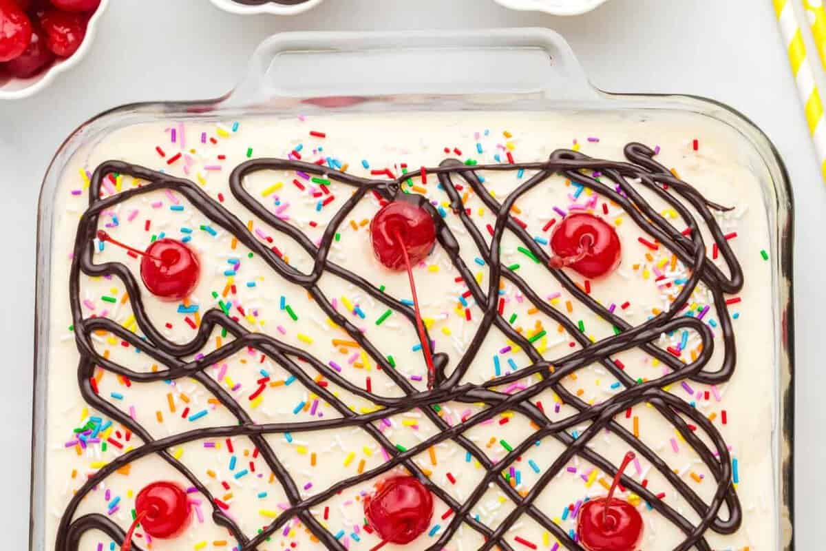 Ice cream sundae cake in a rectangle baking pan, decorated with hot fudge, sprinkles, and cherries.