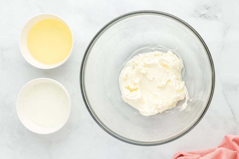 Beaten cream cheese in a glass mixing bowl surrounded by ingredients.
