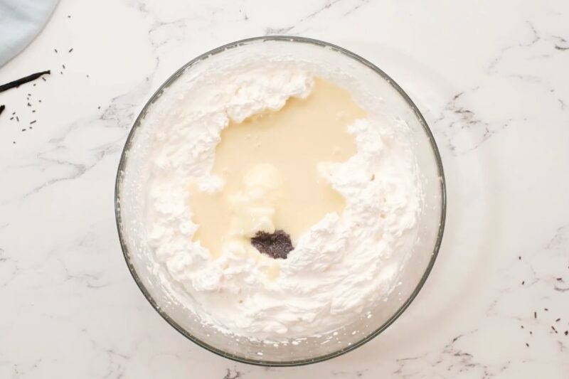 Sweetened condensed milk and vanilla bean paste added to a bowl of whipped cream.