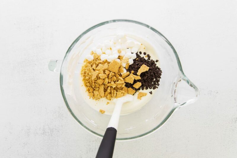 Folding crushed graham crackers, marshmallows, and chocolate chips into cream mixture.