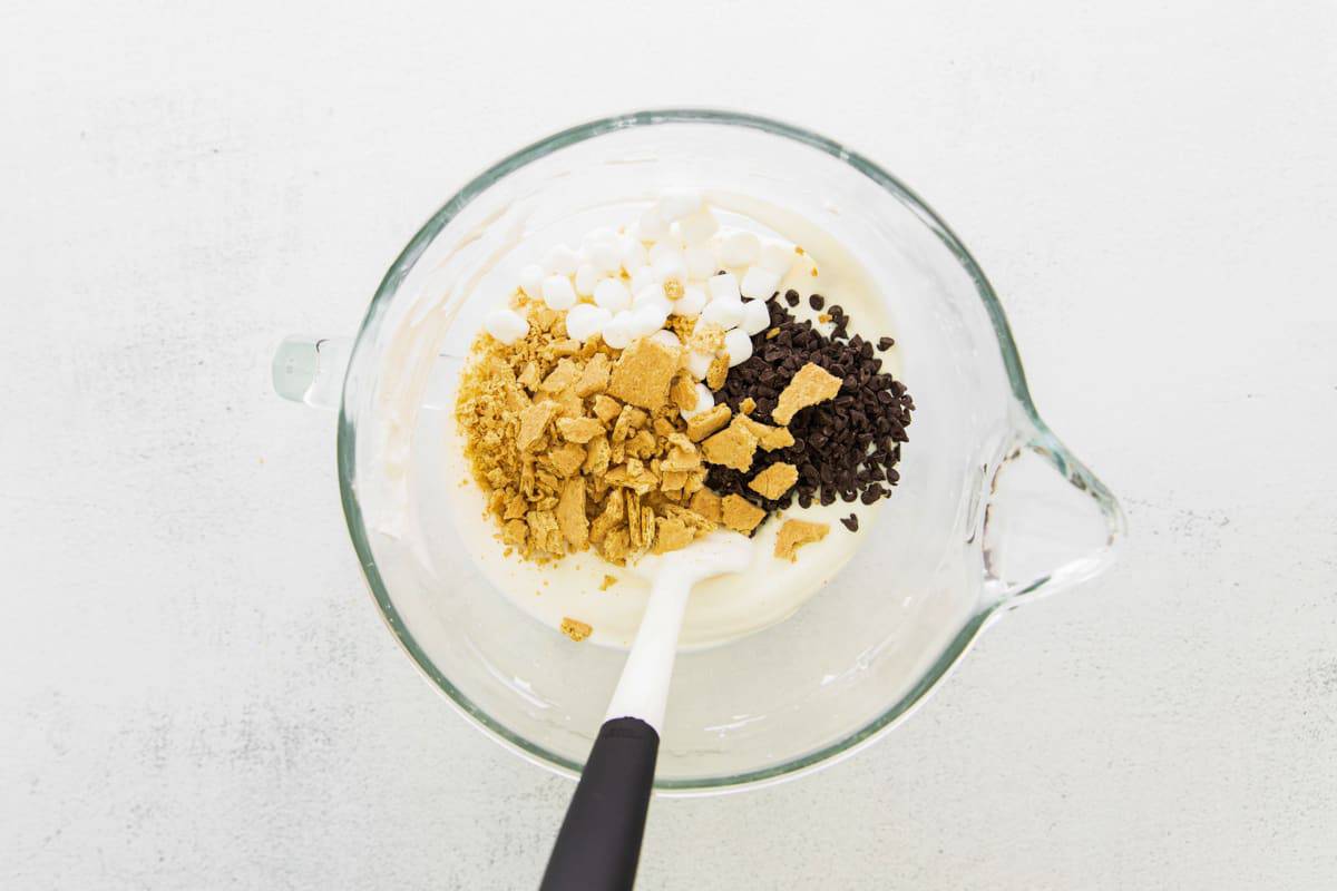 Folding crushed graham crackers, marshmallows, and chocolate chips into cream mixture.