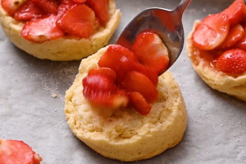 Spooning macerated strawberries onto half of a baked biscuit.