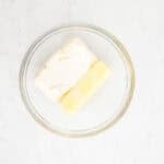 A block of cream cheese and a stick of butter in a glass mixing bowl.