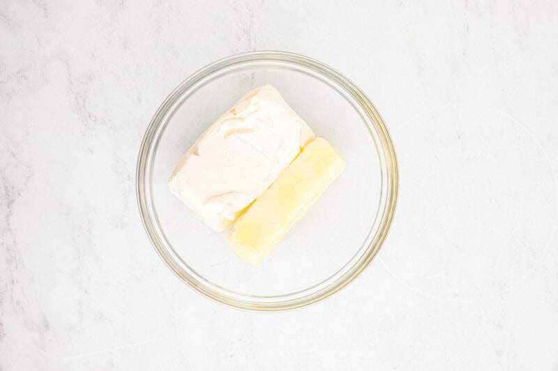 A block of cream cheese and a stick of butter in a glass mixing bowl.