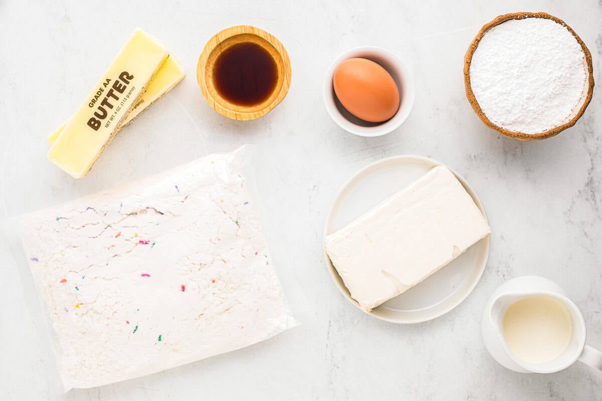 Ingredients for unicorn bars, including funfetti cake mix, one egg, two sticks of butter, heavy cream, cream cheese, powdered sugar, and vanilla extract.