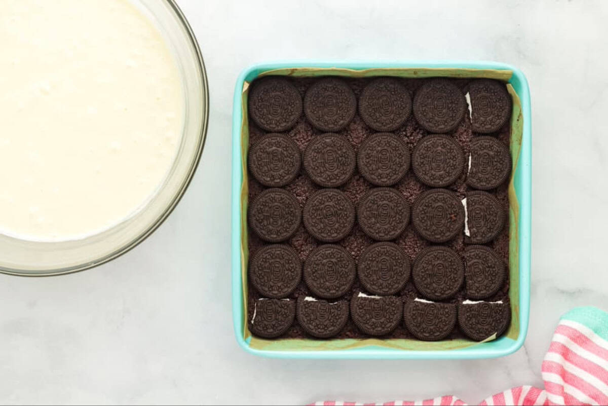 Chocolate cookie crust topped with a layer of Oreo cookies, next to a bowl of cheesecake batter.