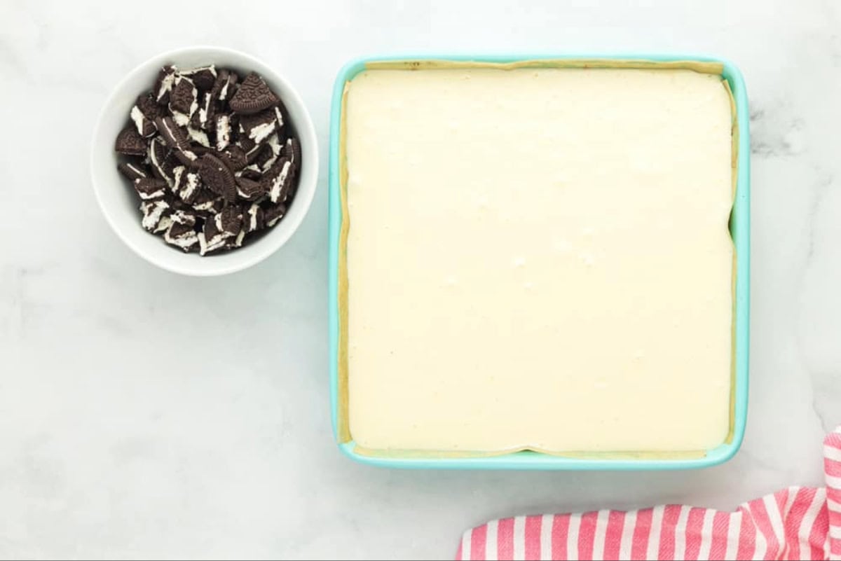 Cheesecake in an 8x8 pan, next to a bowl of crushed Oreos.